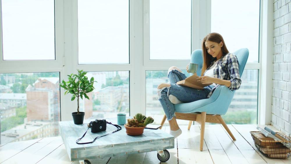 Woman reading a book in a blue armchair by a large window, with plants and a camera on a coffee table.