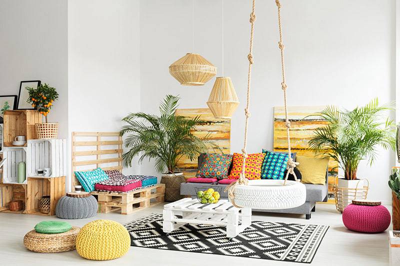 Bright, eclectic living room with colourful cushions, wooden pallet furniture, hanging lamps, and indoor plants.