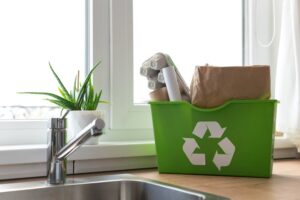 A green recycling bin filled with paper and cardboard by a kitchen sink, with a potted plant nearby.