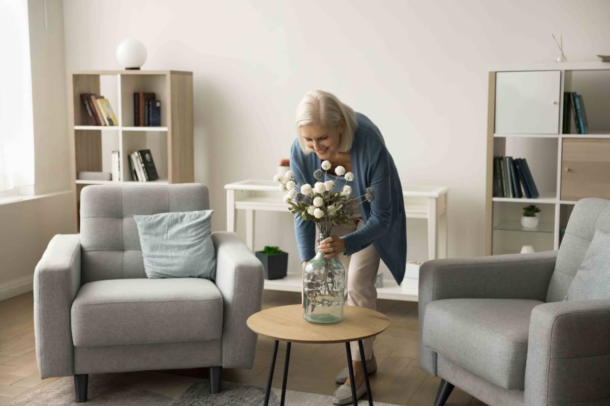 Elderly woman arranging flowers in a vase on a table in a well-lit living room.