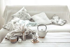 A cosy living room setup with a couch covered in textured pillows and throws, alongside a teapot and dried flowers on a wooden table.