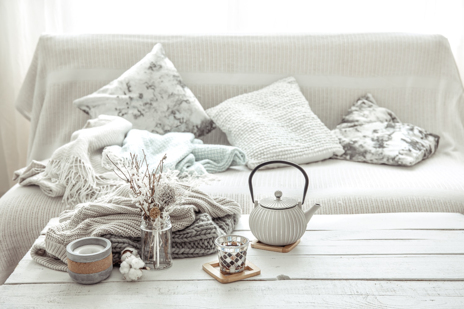 A cosy living room setup with a couch covered in textured pillows and throws, alongside a teapot and dried flowers on a wooden table.