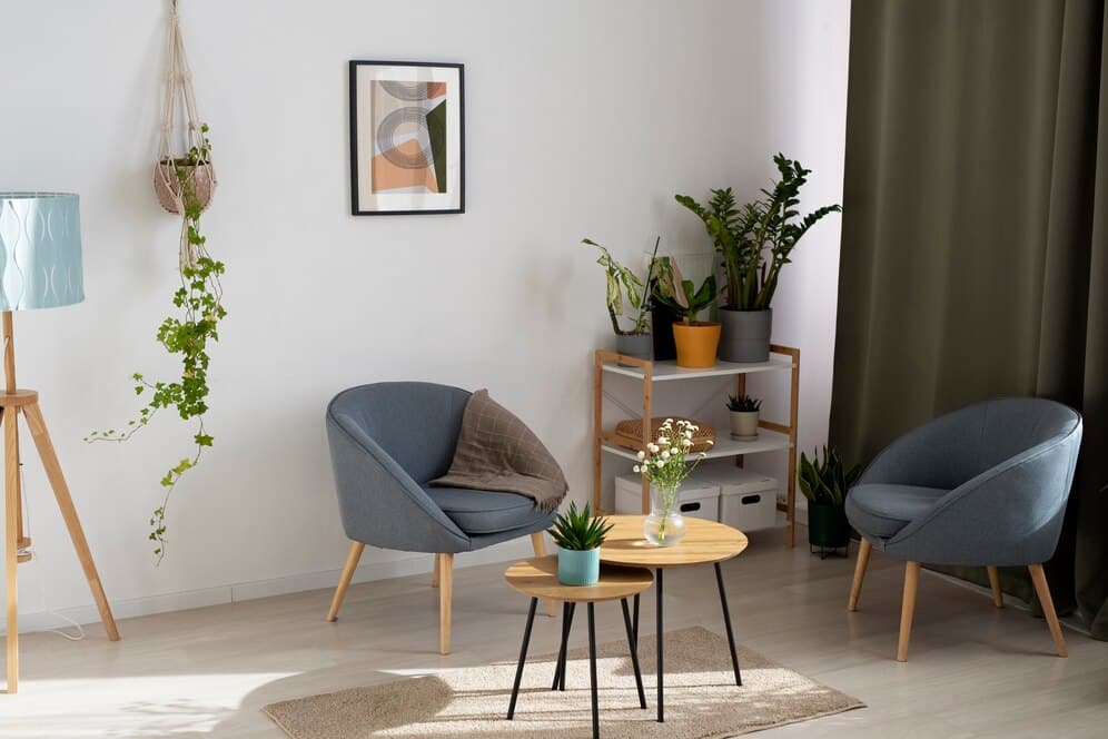 A cozy living room corner featuring modern blue chairs, potted plants, a wooden table, and a decorative lamp by a curtain.