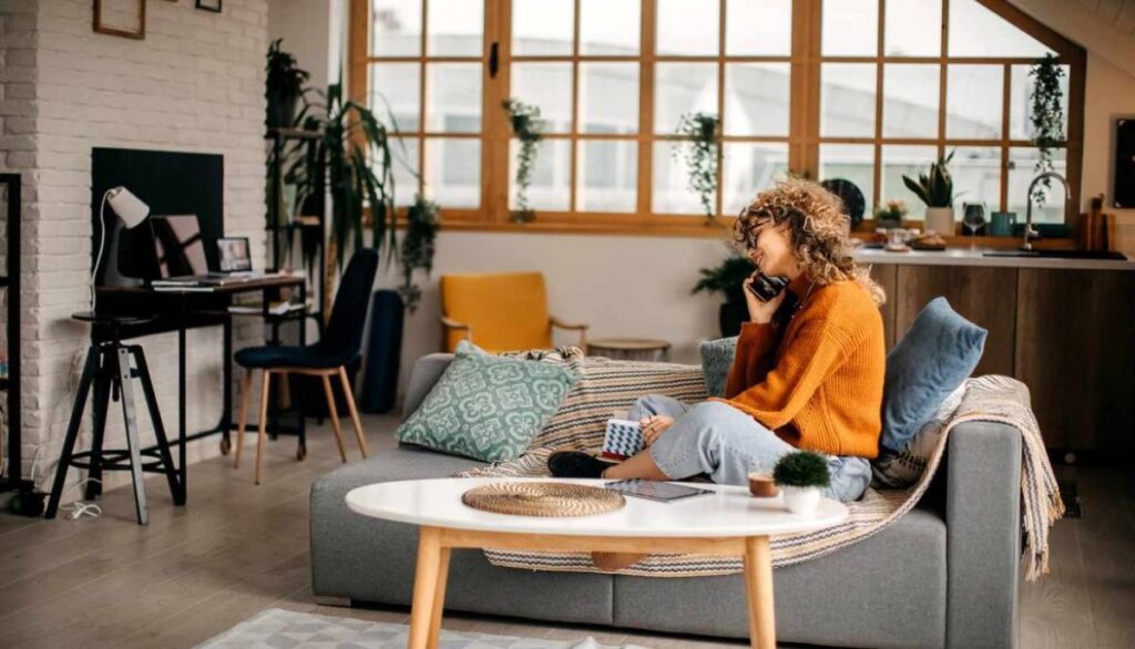 Young woman in an orange sweater laughs on the phone, sitting comfortably on a sofa in a stylish living room.