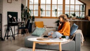 Young woman in an orange sweater laughs on the phone, sitting comfortably on a sofa in a stylish living room.
