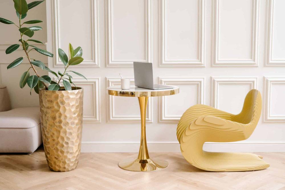 Modern home office with a decorative plant, gold round table, laptop, and a uniquely styled yellow chair against a paneled wall.