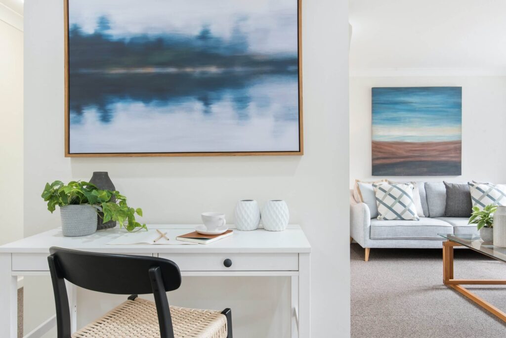 Modern home office desk with a chair and abstract wall art, leading into a cosy living room area with sofa and cushions.