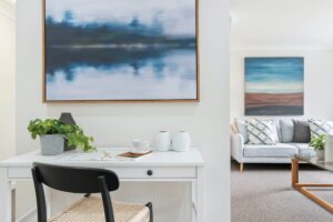 Modern home office desk with a chair and abstract wall art, leading into a cosy living room area with sofa and cushions.