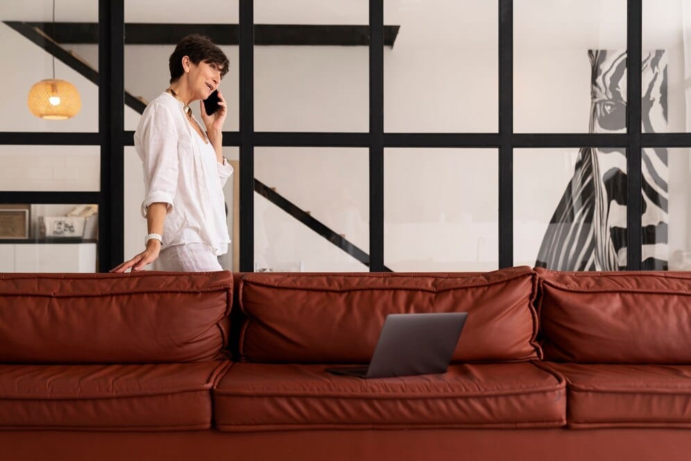 Woman in white shirt talking on mobile phone while walking near a brown sofa in a modern office with large windows and minimalistic decor.