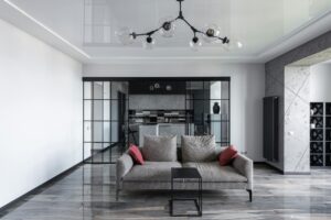 Modern living room with grey sofa, glass pendant lights, and a view into a stylish kitchen behind glass partitions.
