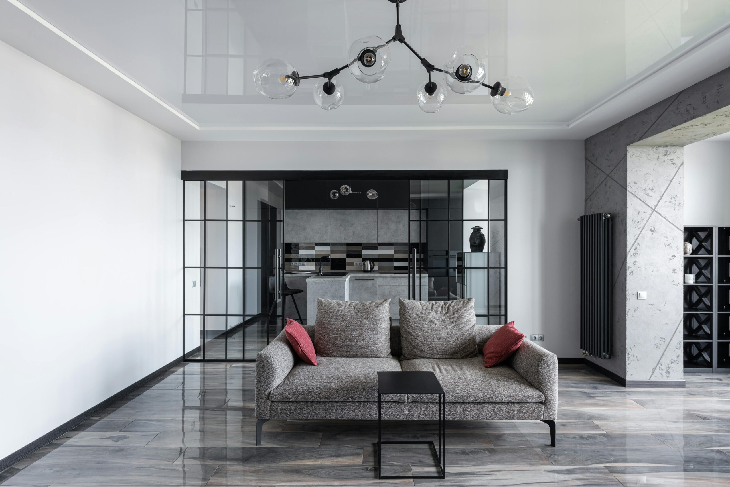 Modern living room with grey sofa, glass pendant lights, and a view into a stylish kitchen behind glass partitions.