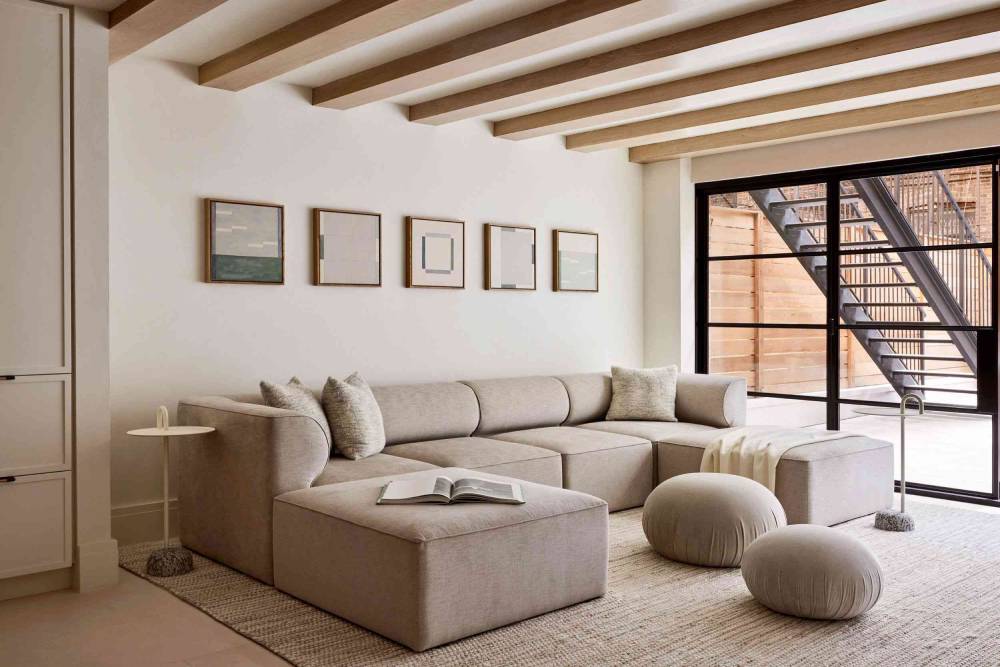  Modern living room with neutral-toned sectional sofa, beams on ceiling, and view of a staircase through glass doors.
