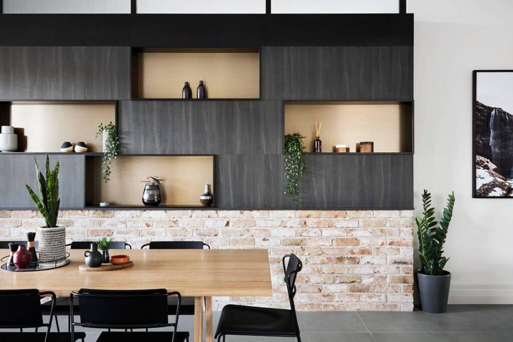 Modern dining area with a wooden table, black chairs, and a shelving unit against a brick wall, decorated with plants and ceramics.