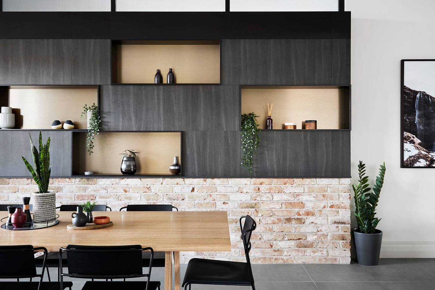 Modern dining area with a wooden table, black chairs, and a shelving unit against a brick wall, decorated with plants and ceramics.