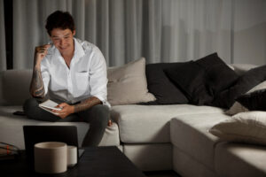 Man with tattoos sitting on a couch, writing in a notebook, smiling during a phone call.