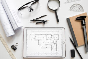 Architectural plans displayed on a tablet and paper surrounded by drafting tools and eyeglasses on a white desk.