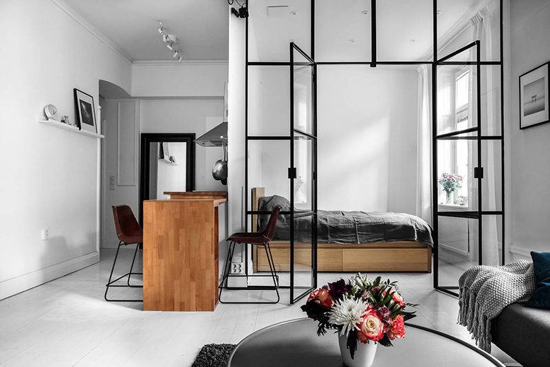  Modern studio apartment with a black framed glass partition separating the bedroom and living area, featuring minimalist furniture and neutral tones.