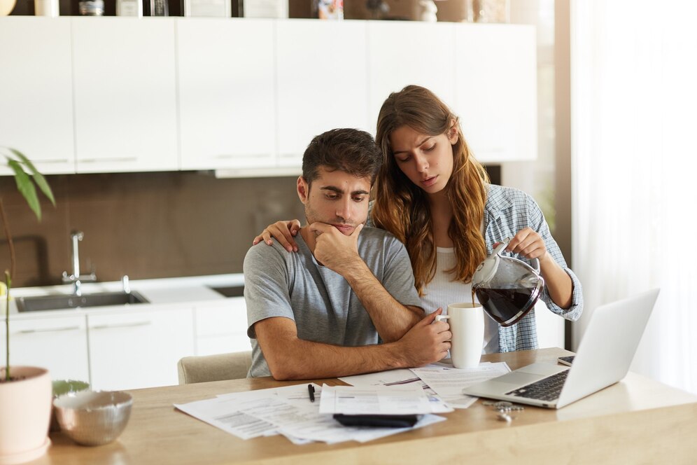 A woman pours coffee while a man looks at a laptop in a modern kitchen.