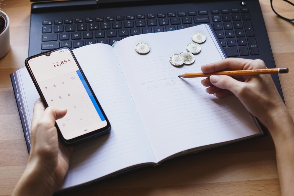 Person using a calculator on a smartphone, writing in a notebook beside coins and laptop on desk.