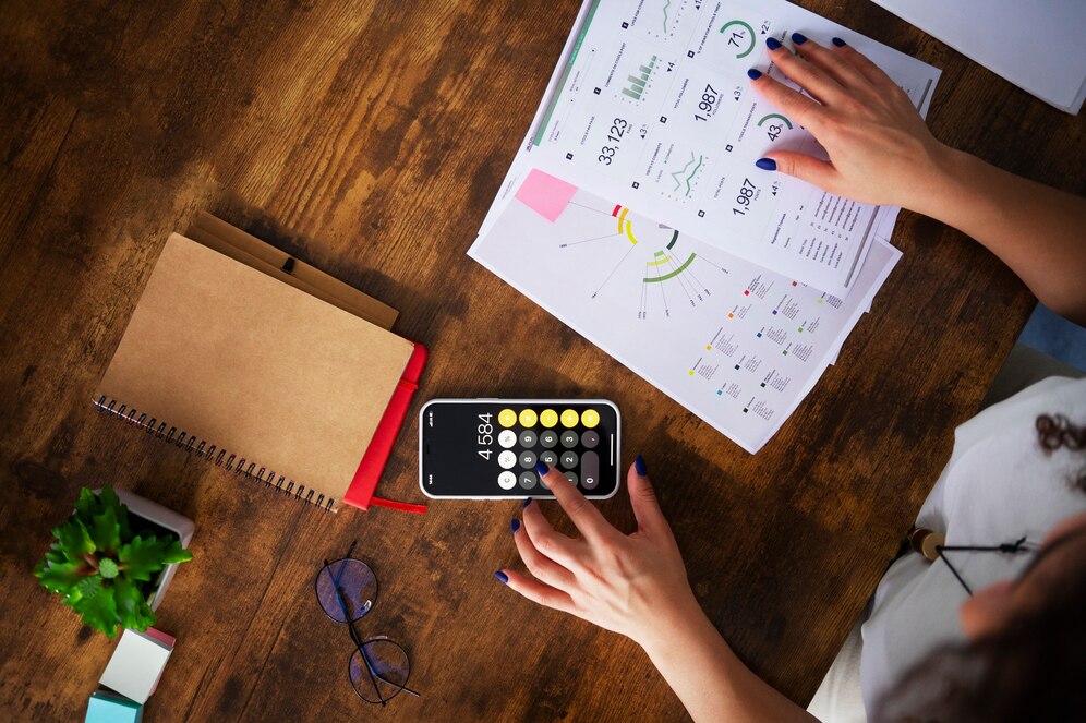 A person calculates data on a smartphone surrounded by financial reports, a notebook a wooden table.