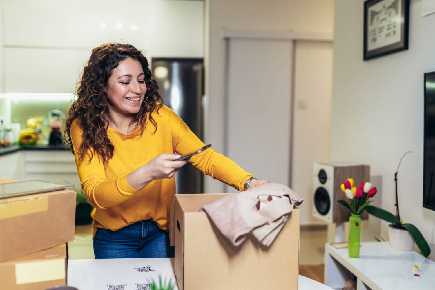 Woman in yellow top unpacking a box in a modern kitchen, smiling as she uses her mobile phone.