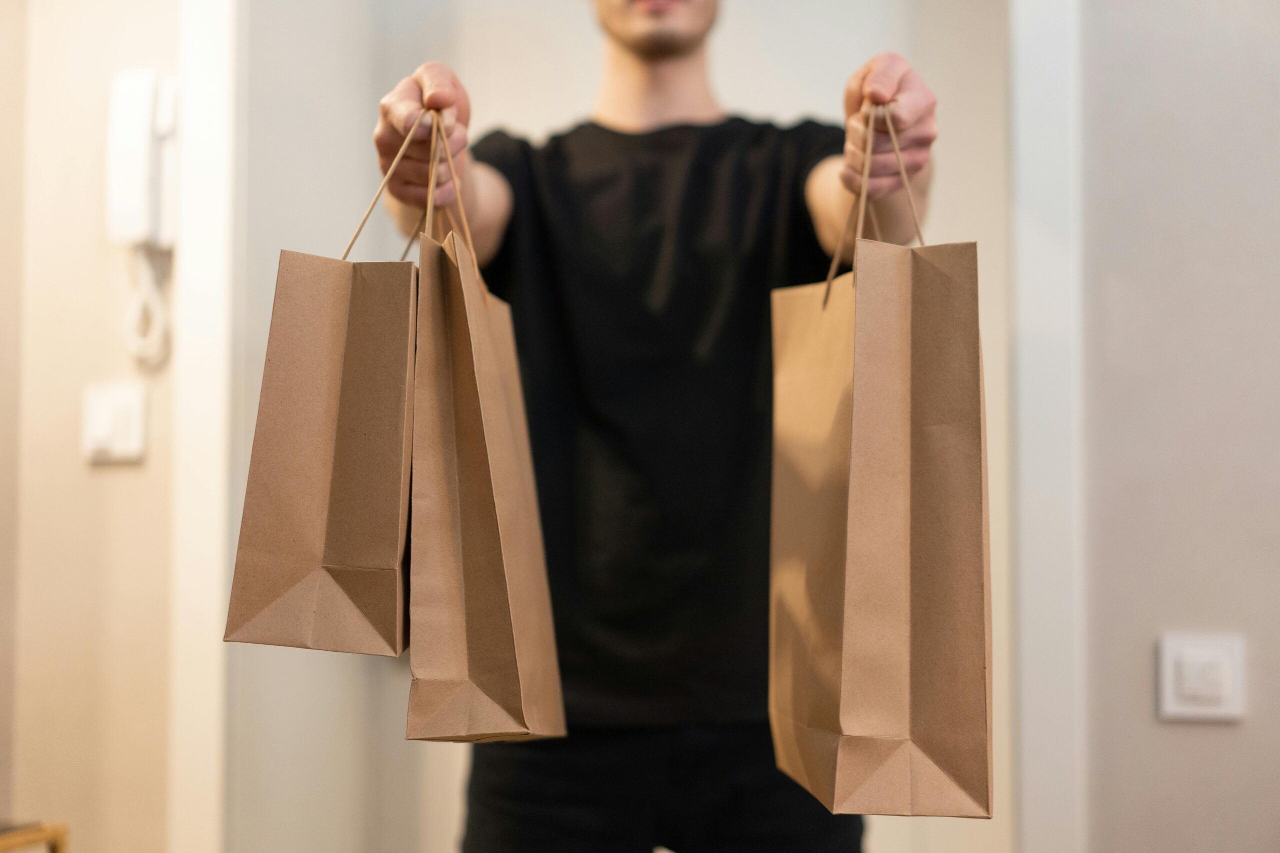 A person in a black shirt holds three brown paper bags, extending them forward in a home setting.