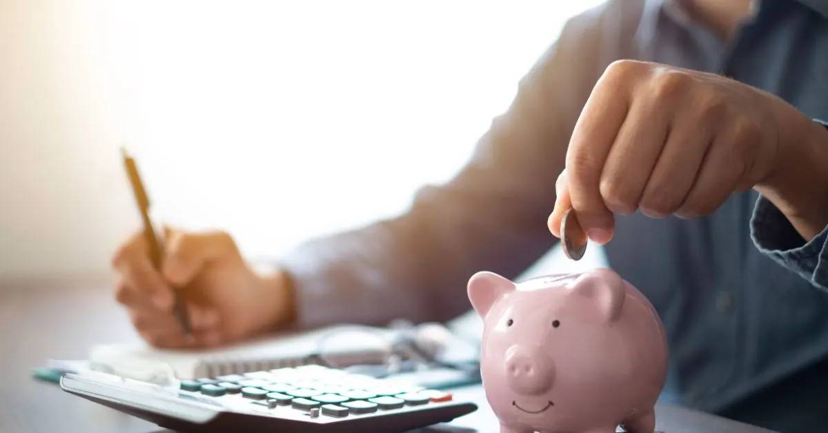 Person inserting coin into a piggy bank while writing notes and using a calculator.