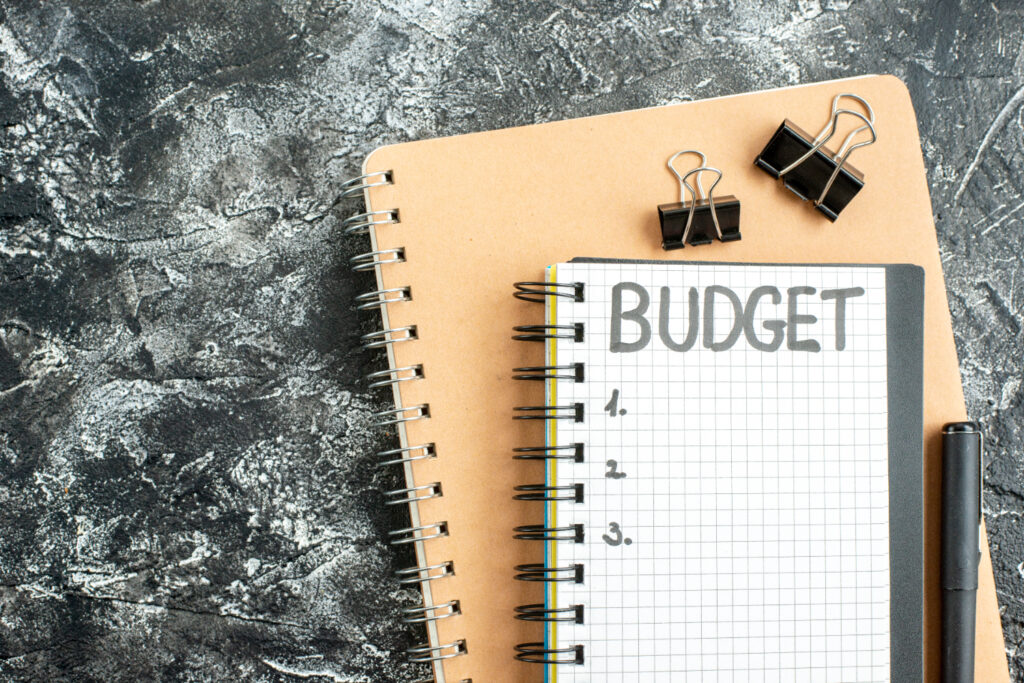 Notebook with BUDGET written on it, pen, and binder clips on a textured surface.