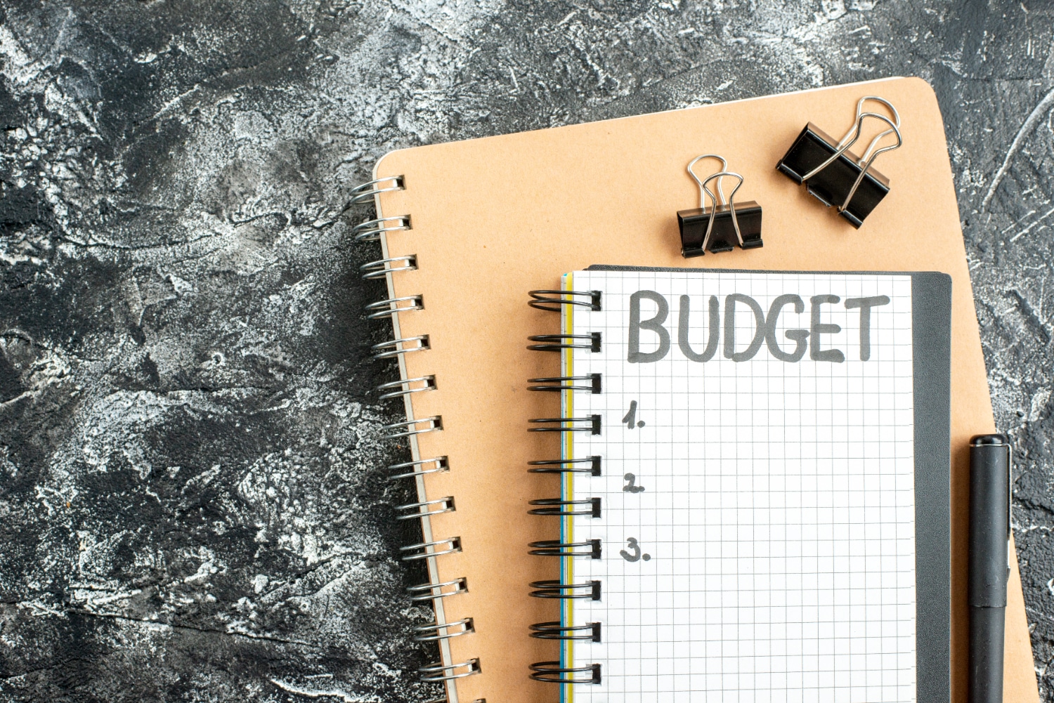 Notebook with BUDGET written on it, pen, and binder clips on a textured surface.