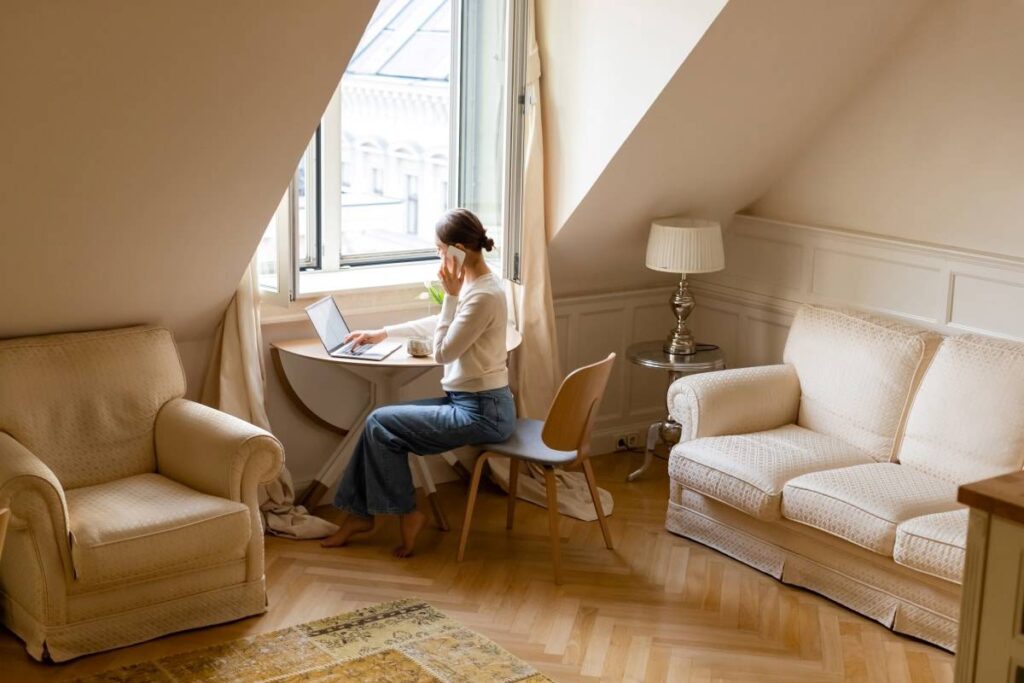 Woman working on a laptop at a small desk by a window in a cozy attic room.