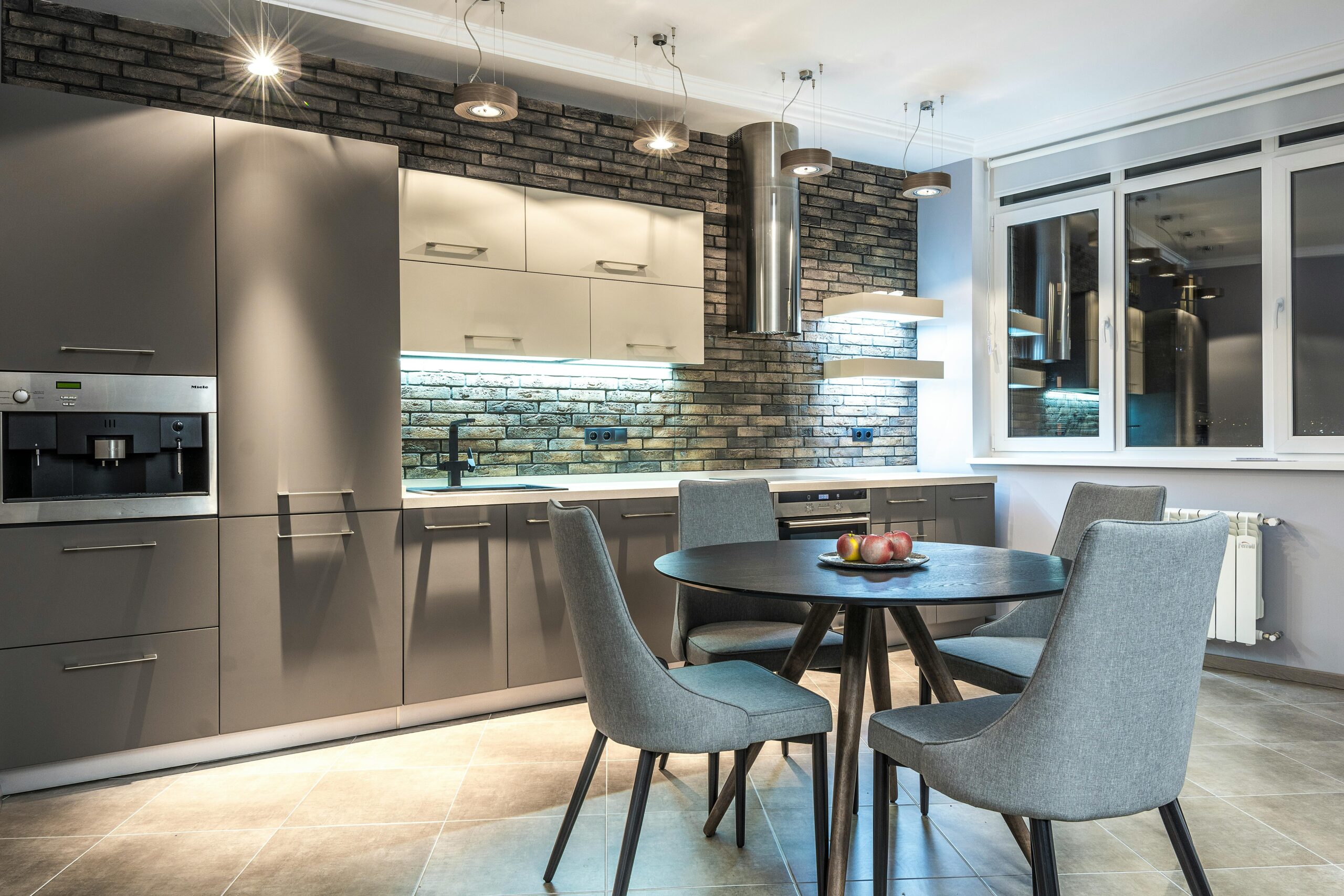 Modern kitchen with grey cabinetry, round dining table, stone backsplash, and sleek pendant lighting.