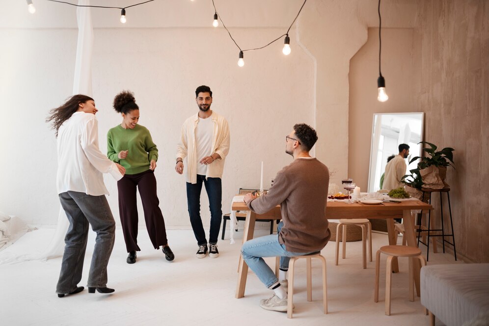 Four adults enjoying a cheerful dance in a cozy, modern living room with another person working at a table.