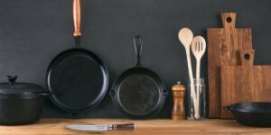 Various cookware and utensils on a wooden surface against a dark background, including cast iron skillets and wooden cutting boards.