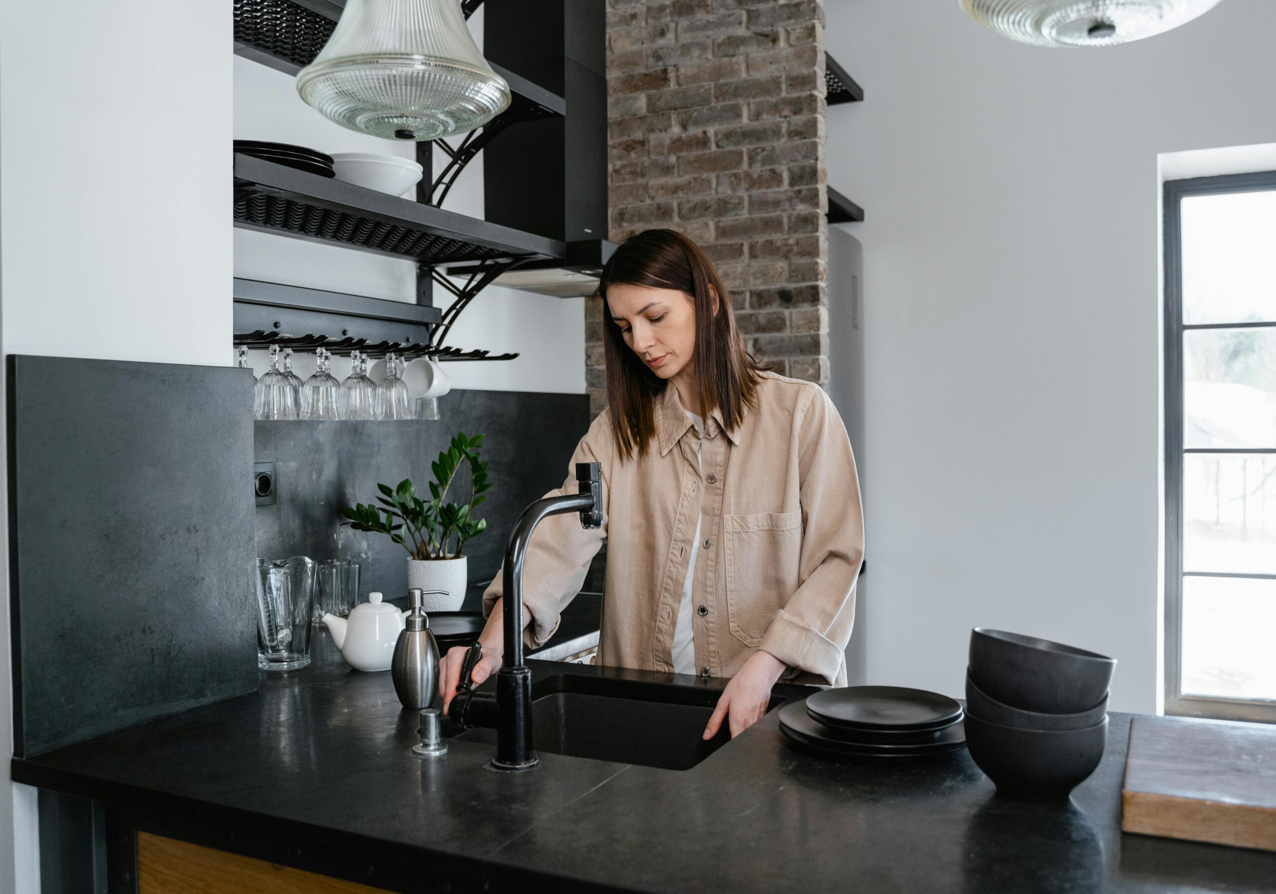 A woman rinses a tray in a modern kitchen with stylish lighting and exposed brick walls.