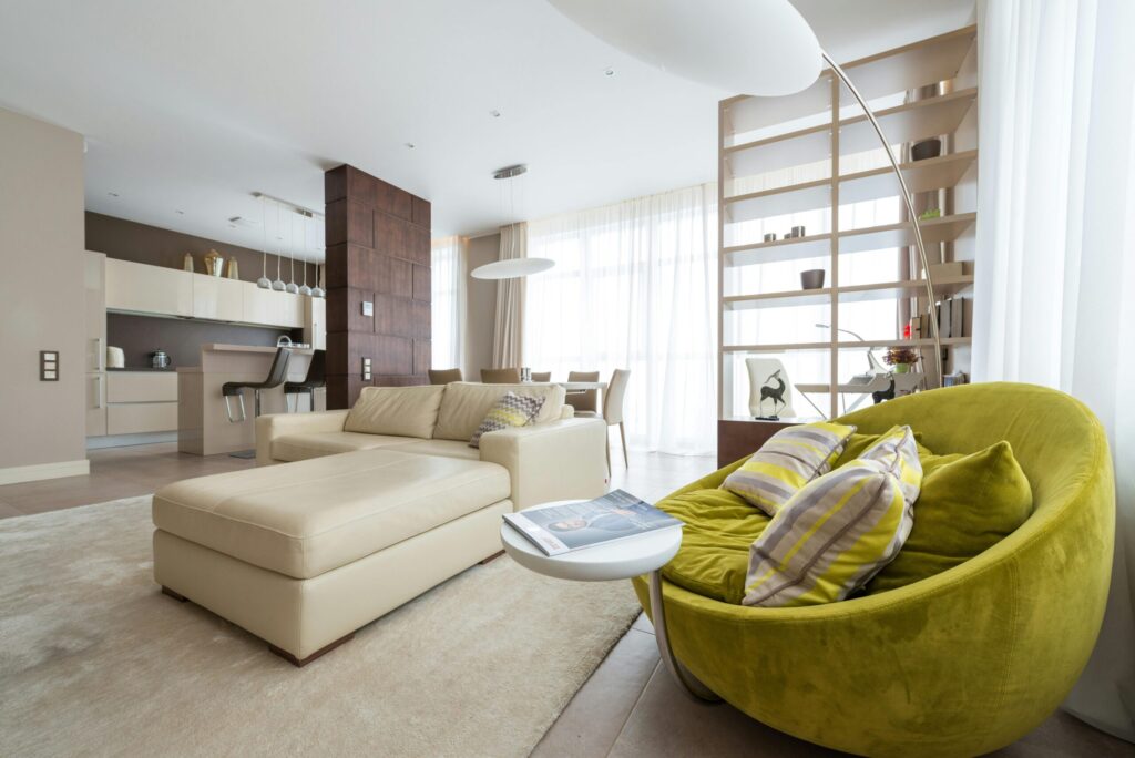 Spacious modern living room with neutral tones and a bright green armchair, large windows, and open kitchen area.