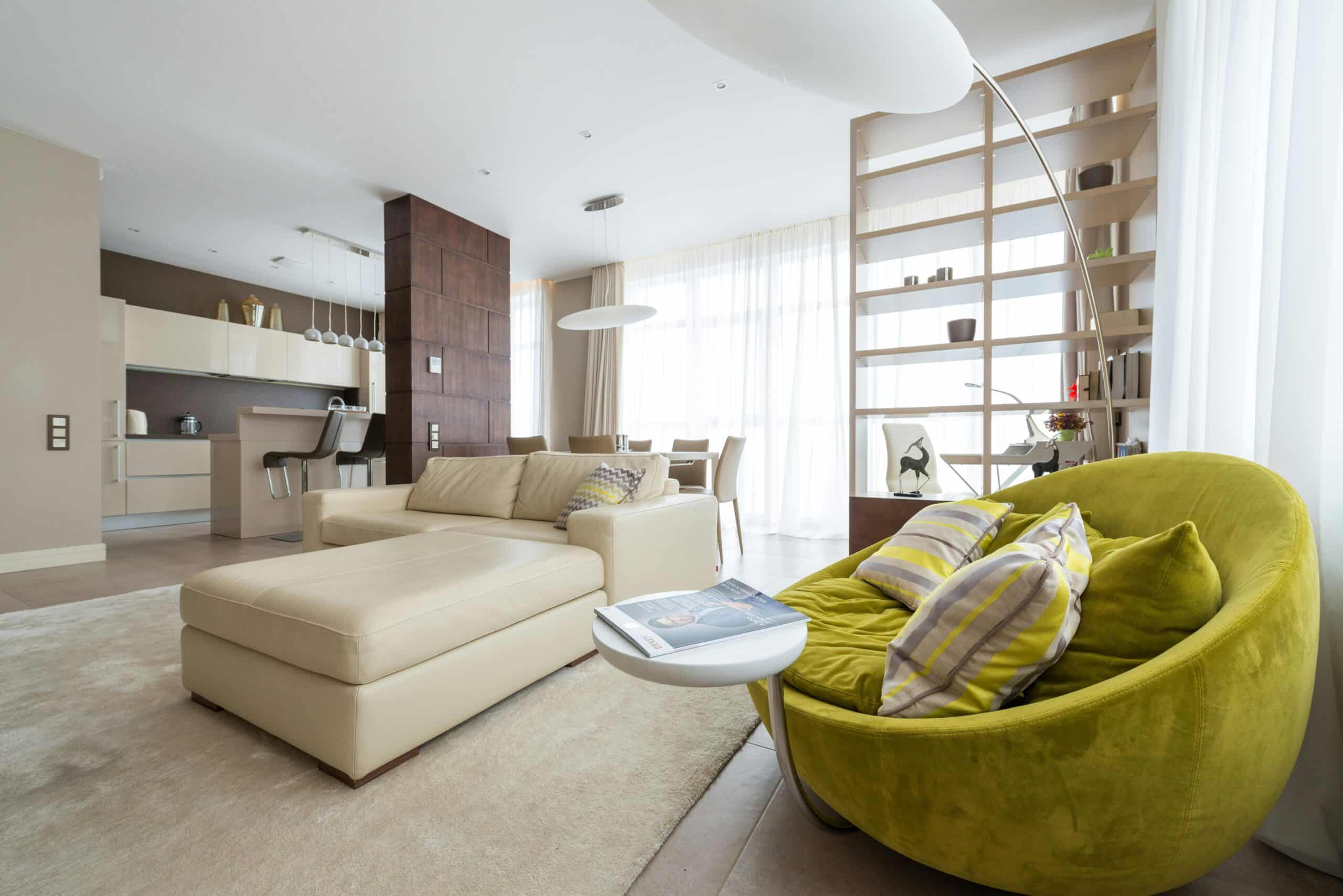 Spacious modern living room with neutral tones and a bright green armchair, large windows, and open kitchen area.