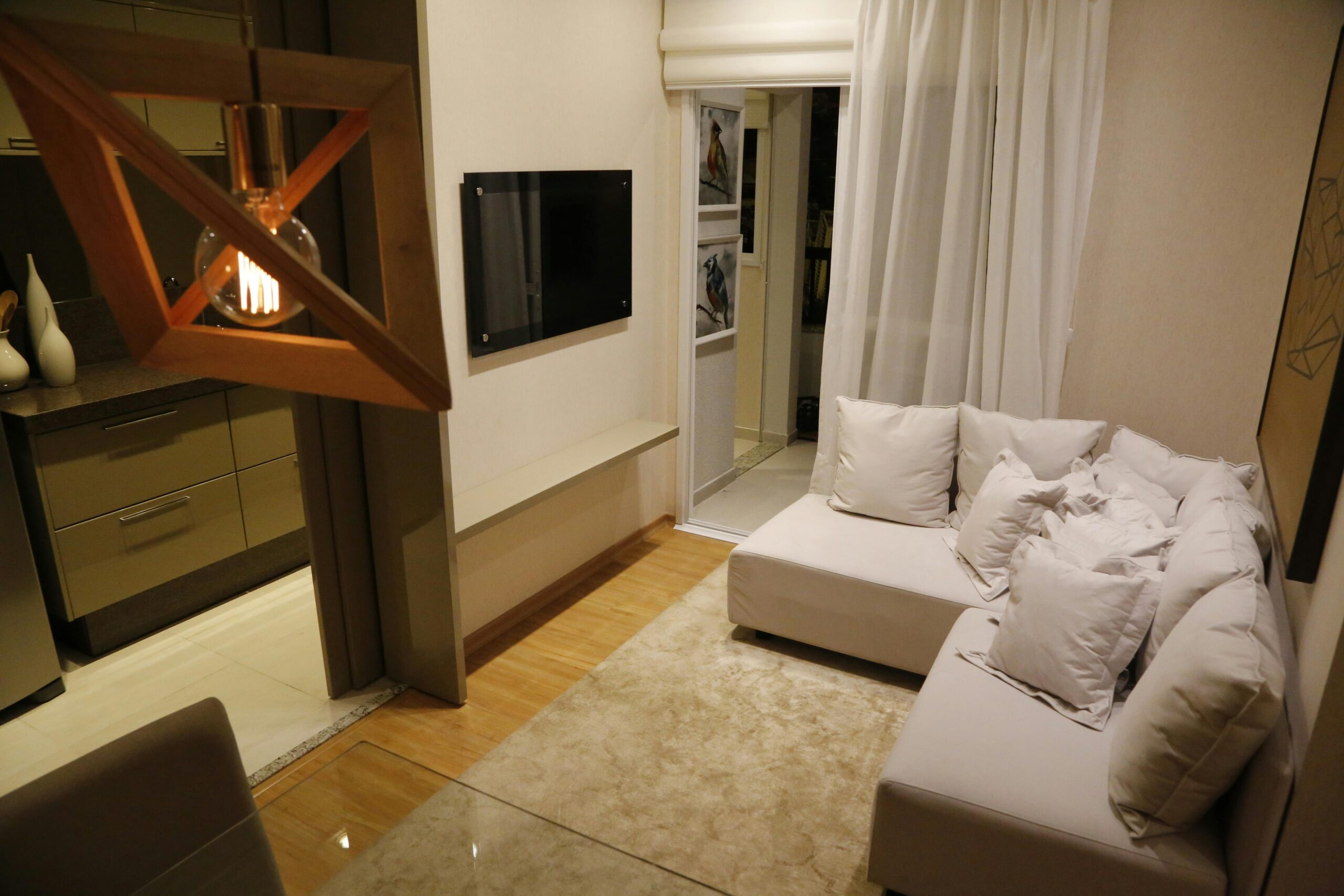 Modern living room with white sofa, decorative pillows, hanging geometric light, and flat-screen TV.