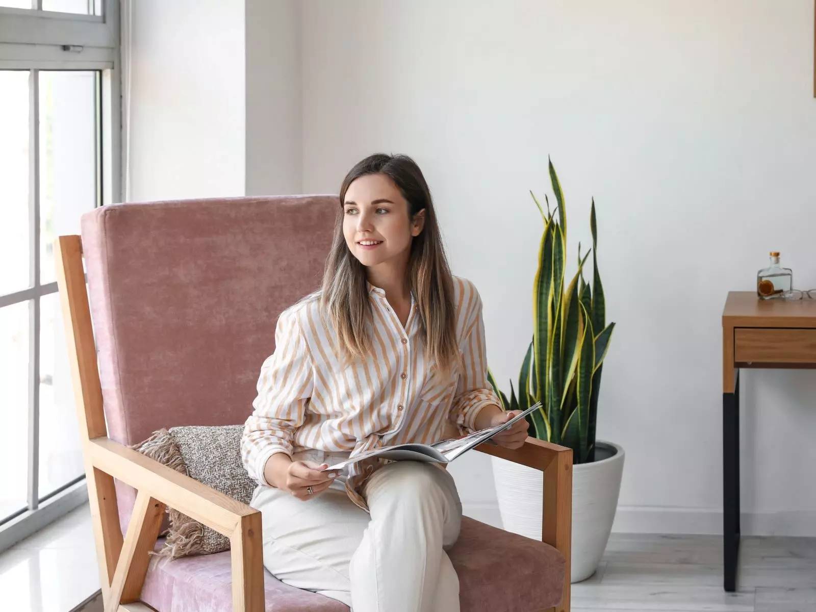 Woman reading a magazine in a modern living room, seated on a pink armchair next to a large potted plant.