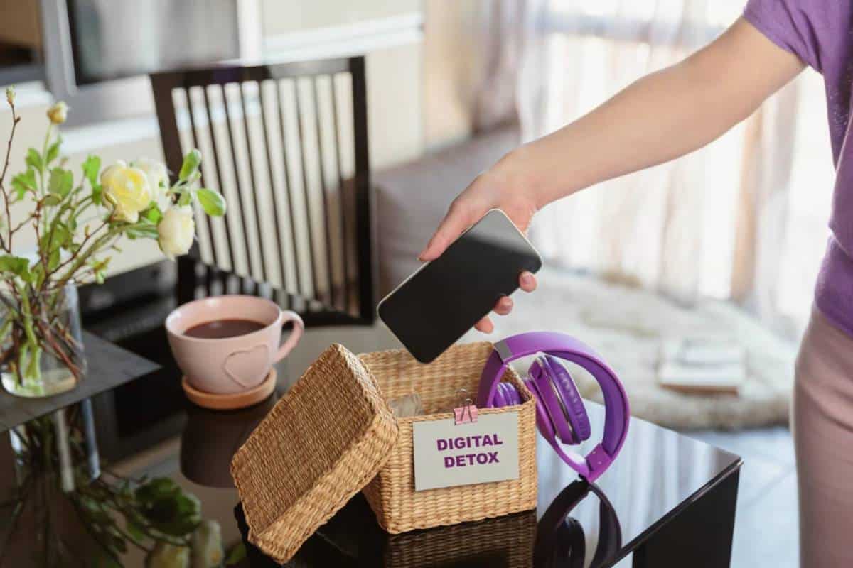 Person placing a smartphone into a basket labelled DIGITAL DETOX beside a pair of headphones on a table.