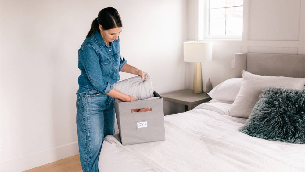  Woman in denim outfit packing a grey storage box on a bed in a neatly arranged bedroom.