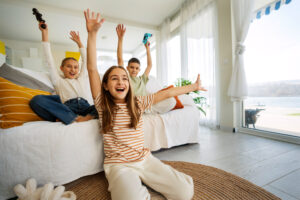 Three children joyfully raising their hands in a bright living room by a large window with a lake view.