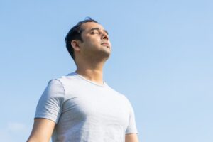 Man in a grey T-shirt standing outdoors, looking upwards with a serene expression against a clear blue sky.
