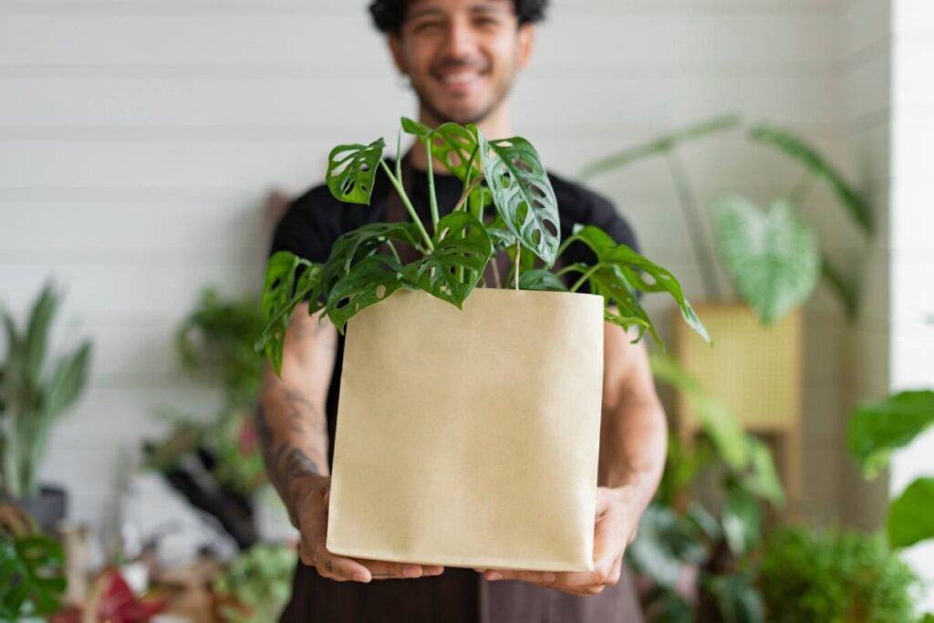 Smiling man holding a monstera plant in a beige pot, surrounded by other greenery.