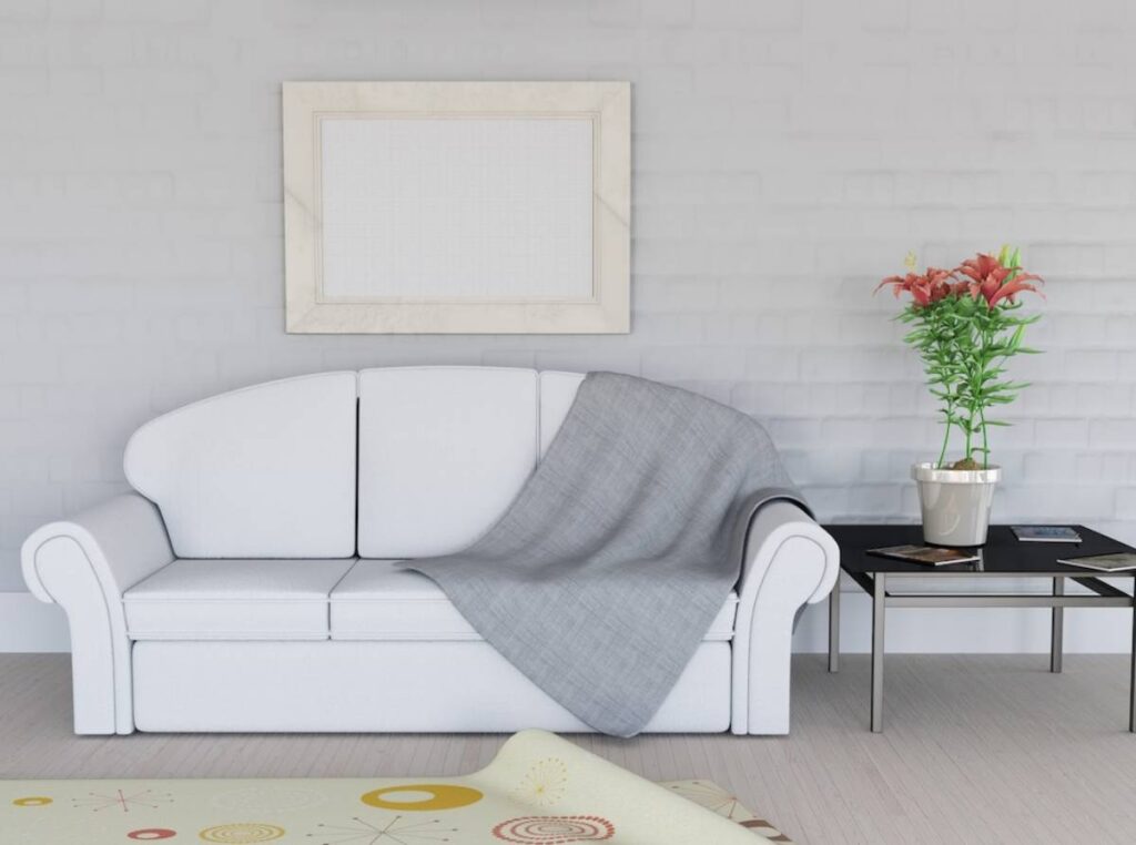 A minimalist living room with a white sofa, grey throw blanket, flowers in a pot, and an empty frame on the wall.