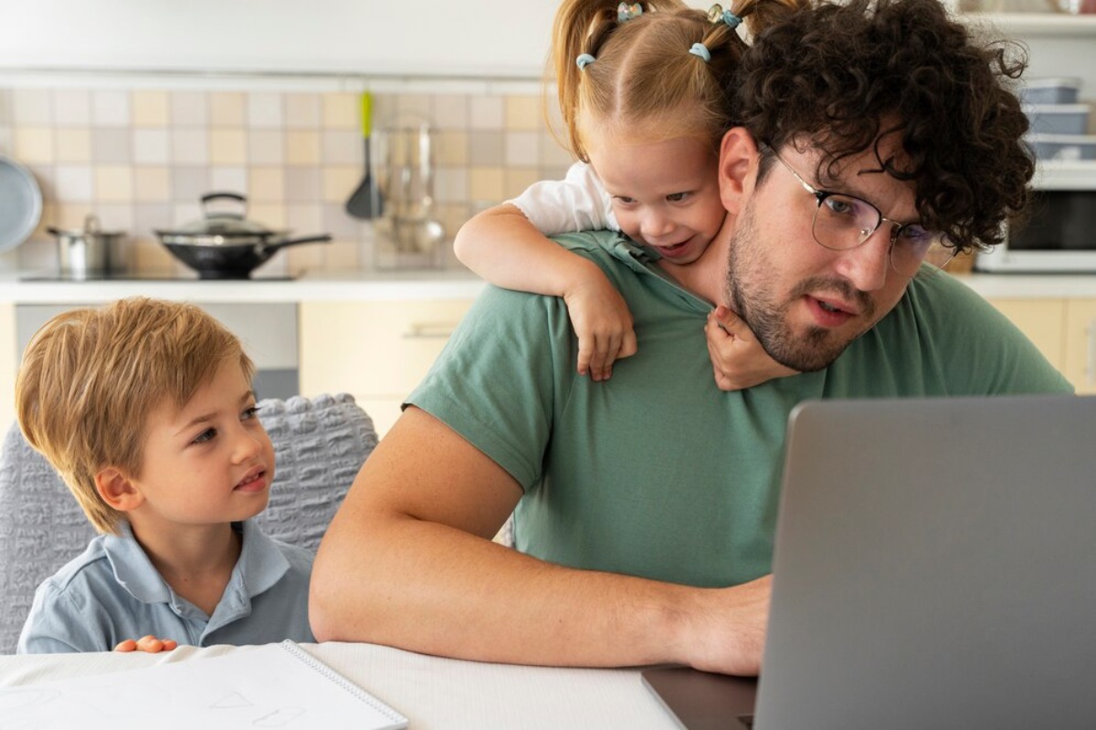 Father with glasses works on laptop, hugged by daughter, while his son sits beside them in a kitchen.