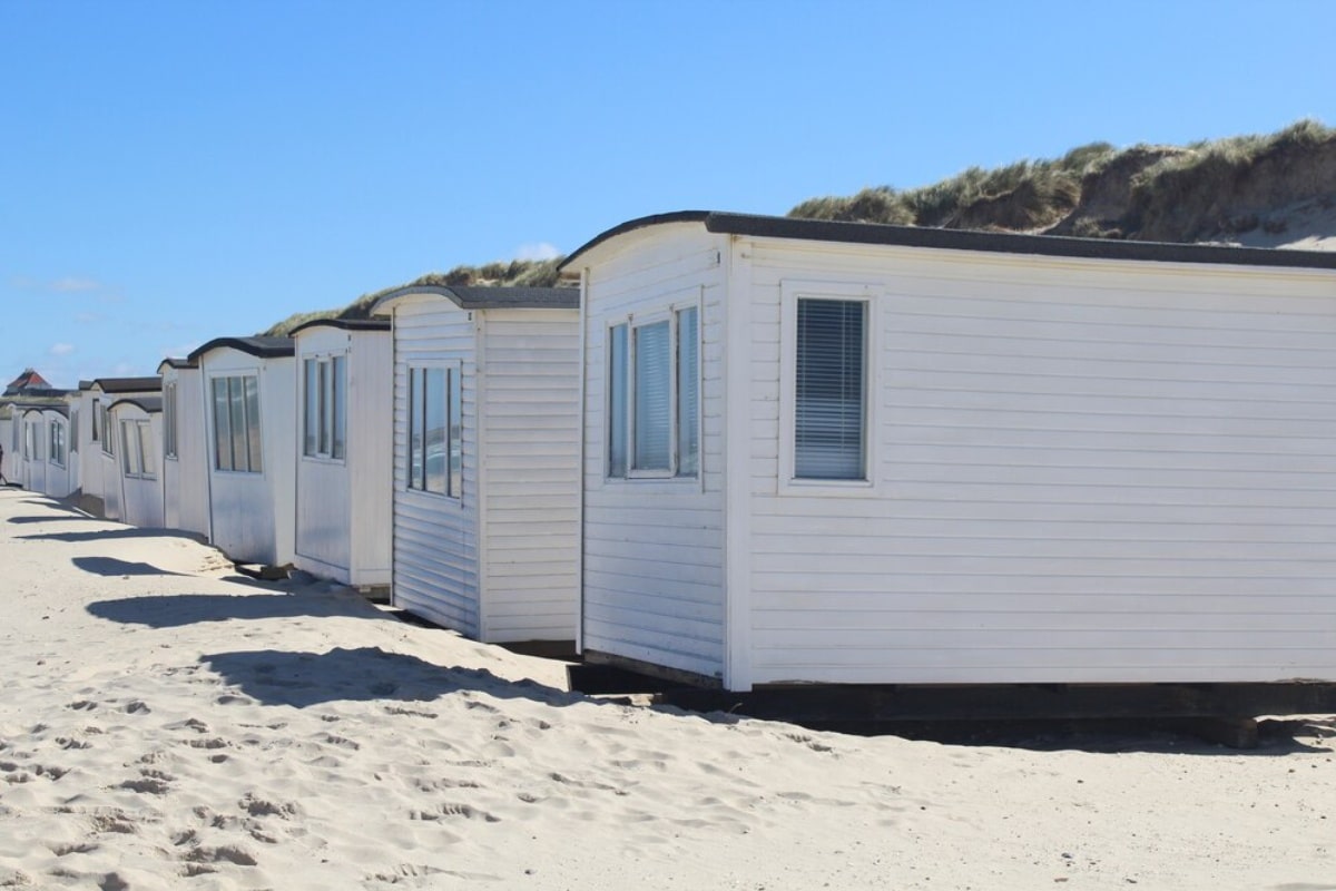Row of white beach huts on sandy shore with dunes in the background under blue sky.