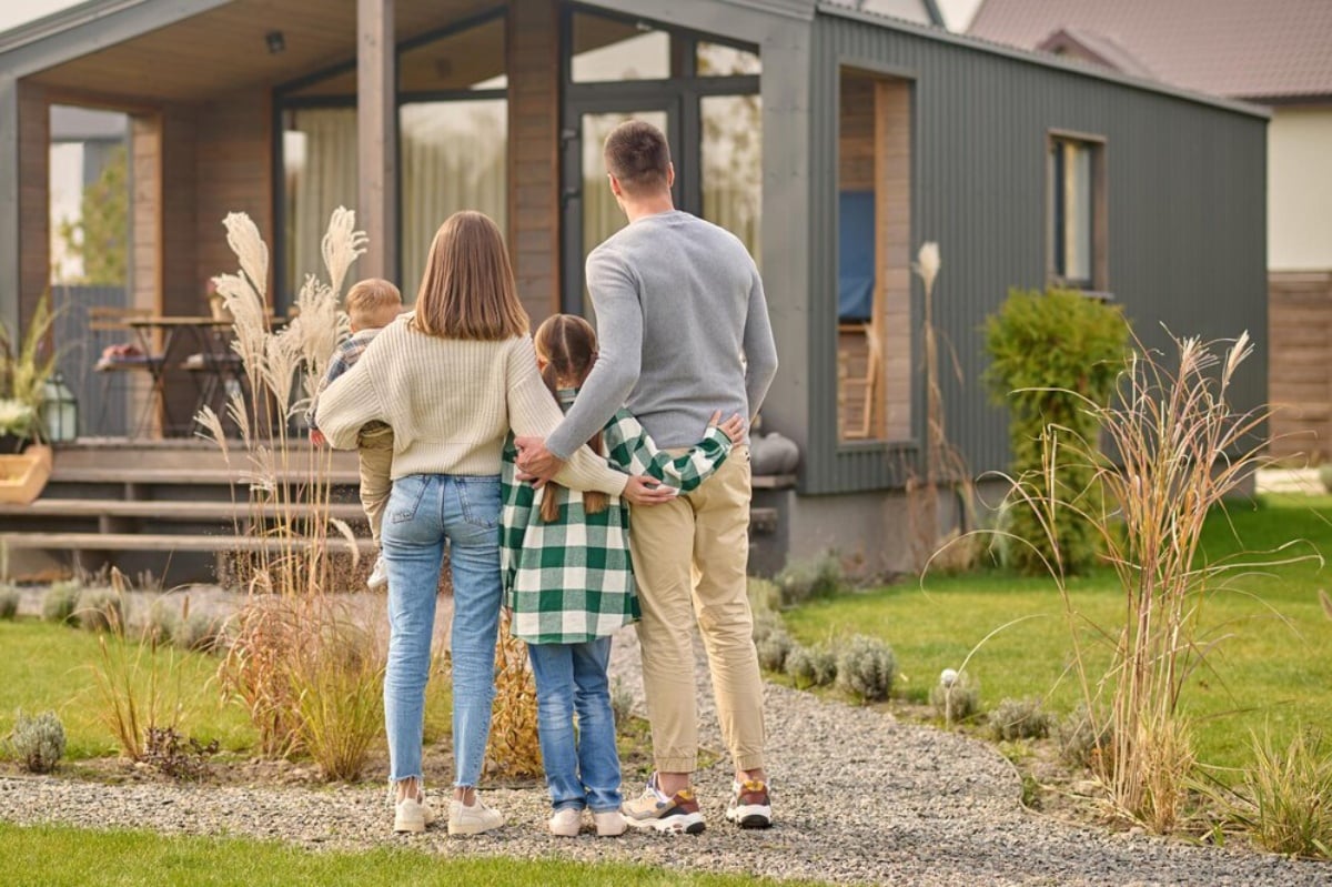 A family of five stands facing a modern house, embracing warmly in their garden.