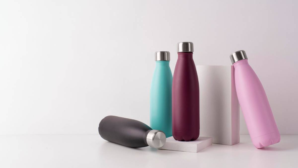Four reusable water bottles in teal, maroon, pink, and dark grey, arranged on and around a white block against a light background. 
