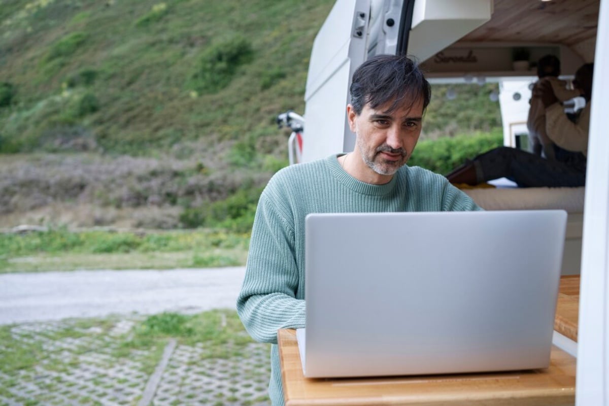 Man using a laptop at a campervan table, with hills in the background.