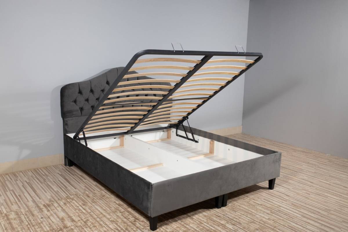 Grey upholstered storage bed with the mattress base lifted to show empty space underneath, set in a room with grey walls. 